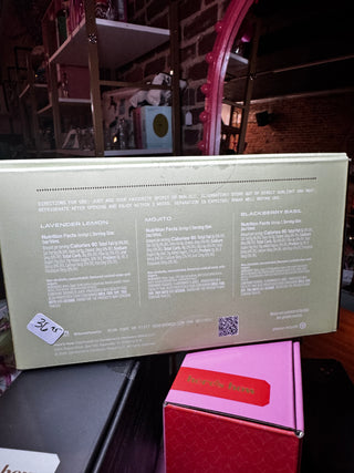 A large pale green rectangular product box with printed ingredient and usage text on the back, placed on top of a pink box in a retail or storage setting. The main visible product is the back of the box, and no person or animal is present.