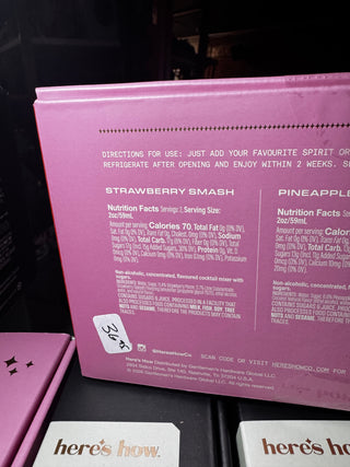 A pink box of Here's How Strawberry Smash cocktail mix, showing the back of the packaging with product details, a white price sticker, and partial neighboring product boxes on a dark surface.