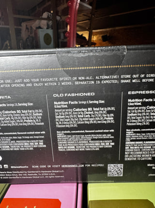 Close-up of the back of a beverage can or box showing printed nutrition facts, flavor labels including Old Fashioned and Espresso, a QR code, and a clear plastic seal strip near the top.