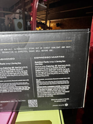 Close-up of the back of a black beverage box showing printed text, nutrition facts, and a QR code. The visible product name reads Espresso Martini, and the box is photographed in a real indoor setting with shelving and other items in the background.