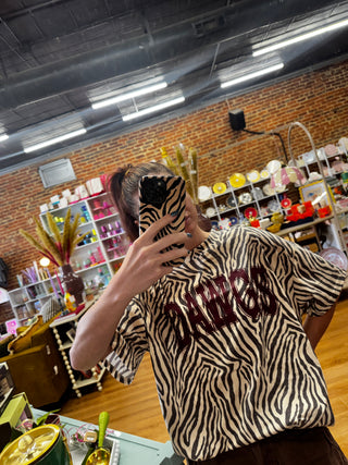 A person taking a mirror selfie in a shop, wearing an oversized short-sleeve t-shirt with a zebra print pattern and dark burgundy collegiate lettering that reads 'DAWGS' across the chest.
