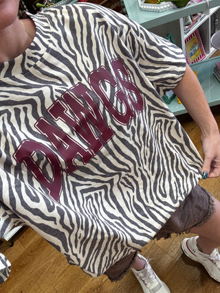 A person wearing an oversized zebra-print t-shirt with 'DAWCS' printed in maroon varsity letters across the chest, paired with brown shorts and white sneakers in a store setting.