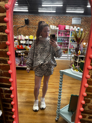 A person taking a mirror selfie in a boutique, wearing a matching zebra-print sweatshirt and shorts set with white sneakers. The boutique has a brick wall, wooden floors, and a large pink-framed mirror.