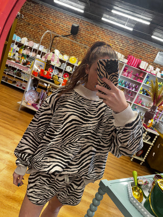 A woman takes a mirror selfie in a boutique while wearing a matching zebra-print lounge set consisting of a long-sleeve sweatshirt and shorts. The background shows shelves filled with colorful items and a brick wall.