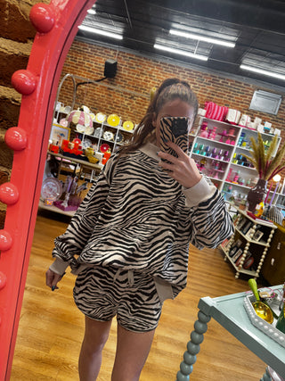 A person taking a mirror selfie in a boutique shop, wearing a matching zebra-print lounge set consisting of a long-sleeve sweatshirt and shorts. The background shows shelves filled with colorful items and a brick wall.
