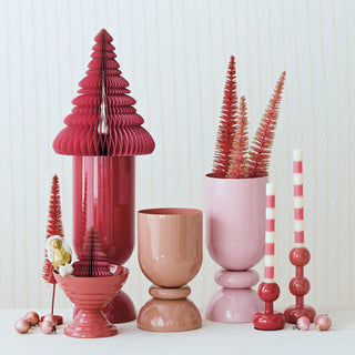 A collection of pink and red holiday decorative items, including a large red accordion Christmas tree, a pink bottle tree, a small gold ornament, striped candlesticks, and ceramic vases, arranged on a white surface against a light textured wall.