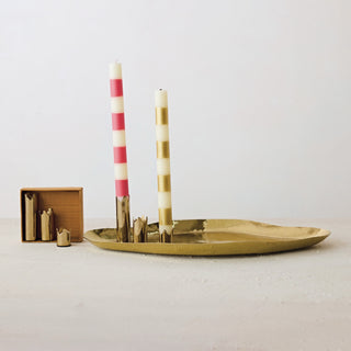 A gold-toned metal candle holder tray with two decorative candles, one pink and white striped and one gold and white striped, displayed on a light surface against a white background.