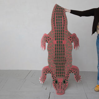 Person holding a large pink alligator-shaped mat against a white wall.