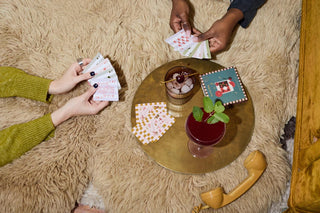 Two people playing cards on a fluffy beige carpet with a small table featuring a cocktail and snacks.