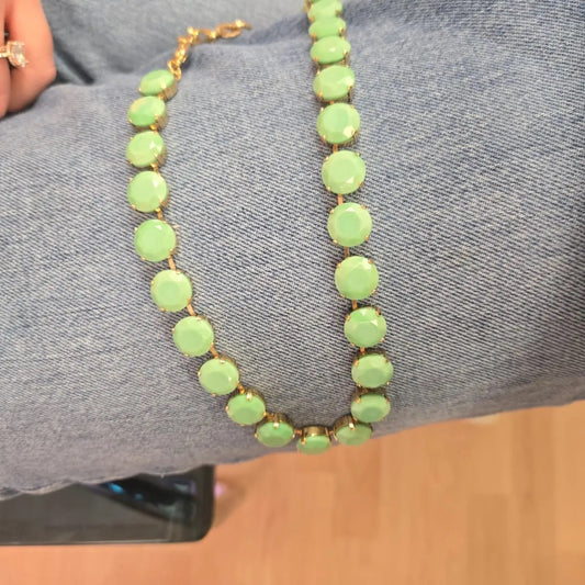 A gold-tone chain necklace with round light green faceted beads draped over blue denim fabric.