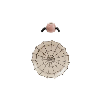 A Halloween spider-themed table setting featuring a spider-shaped pink pot with black legs and a matching spiderweb-patterned plate below it, set against a plain white background.