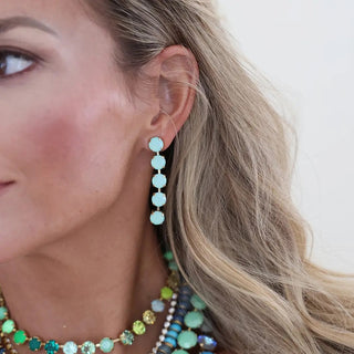 A close-up of a woman's ear wearing a dangling earring made of five light blue-green round beads connected by small gold-toned links. She also wears layered beaded necklaces, but the focus is on the earring.