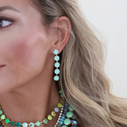 A close-up of a woman's ear wearing a dangling earring made of five light blue-green round beads connected by small gold-toned links. She also wears layered beaded necklaces, but the focus is on the earring.