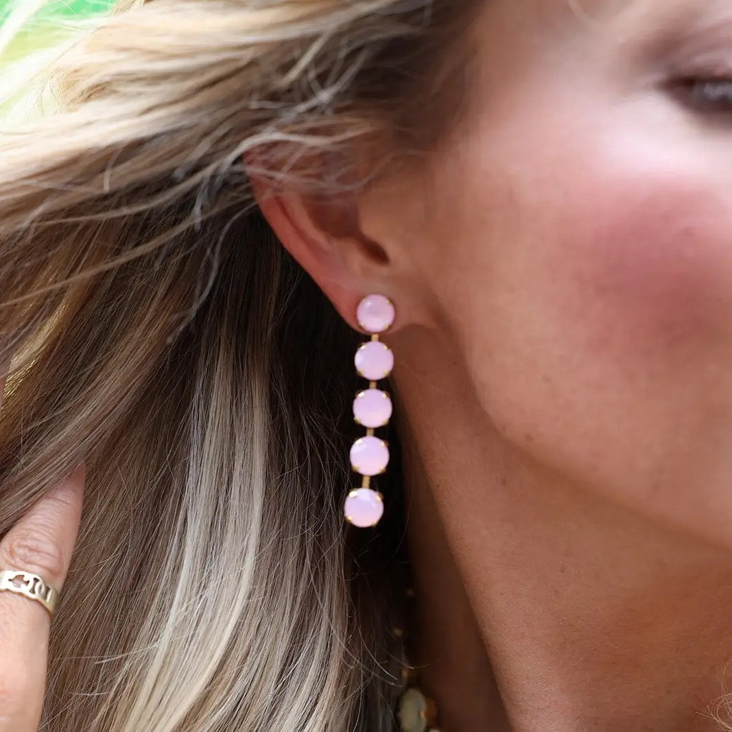 A close-up of a woman wearing a long drop earring with five stacked light pink round stones in gold-toned settings.