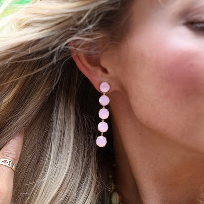 A close-up of a woman wearing a long drop earring with five stacked light pink round stones in gold-toned settings.
