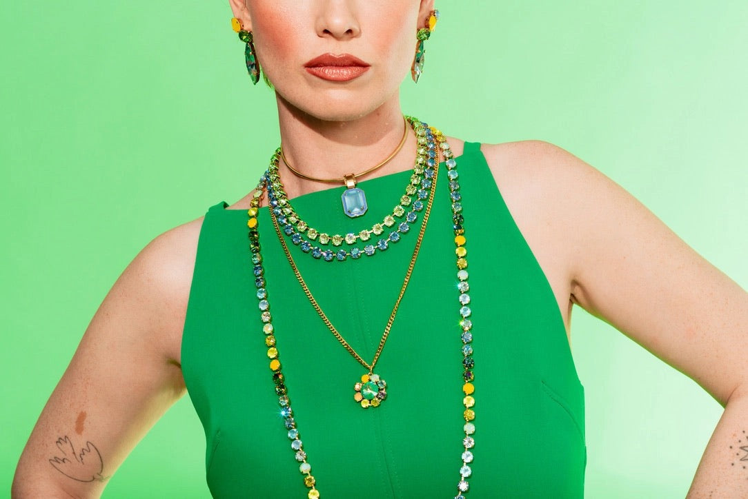A model wearing a vibrant green sleeveless top and layered gold necklaces adorned with green and yellow rhinestones, set against a solid light green background.