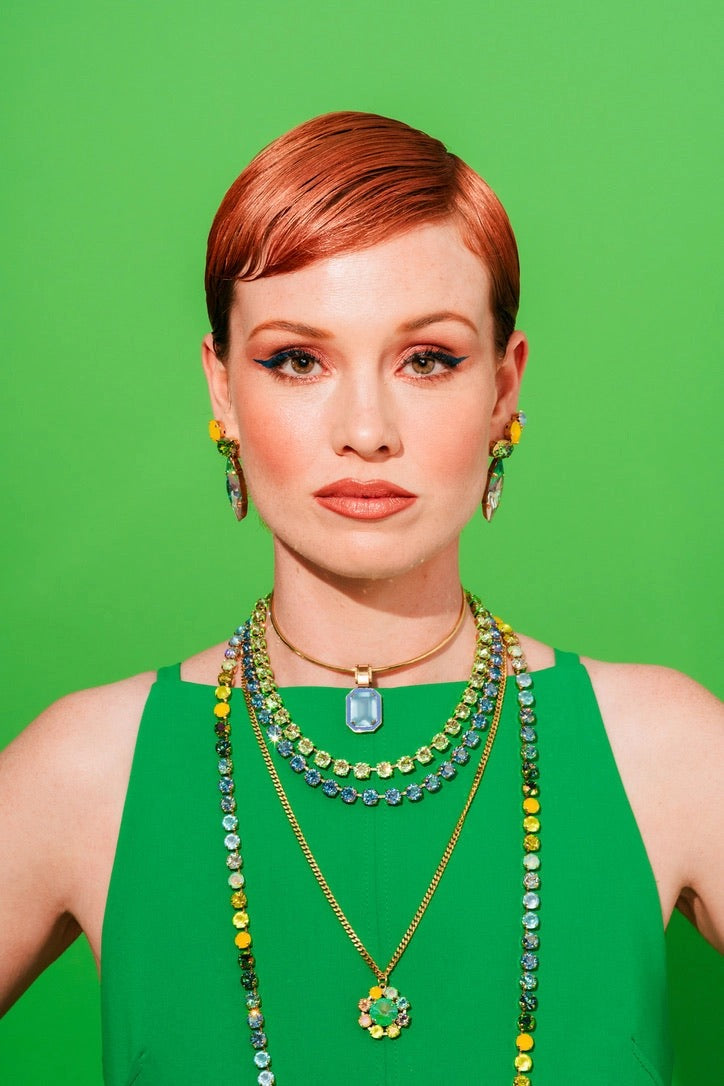 A woman with short red hair wearing a green sleeveless top and layered gold necklaces with green and yellow beaded details, set against a solid green background.
