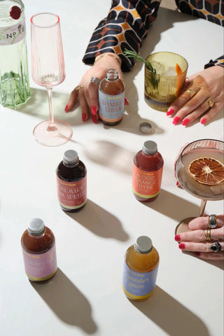 A styled flat-lay of several small bottles of syrup or juice with colorful labels, arranged on a white surface with cocktail glasses and hands holding them. No clear product brand is legible on the bottles.