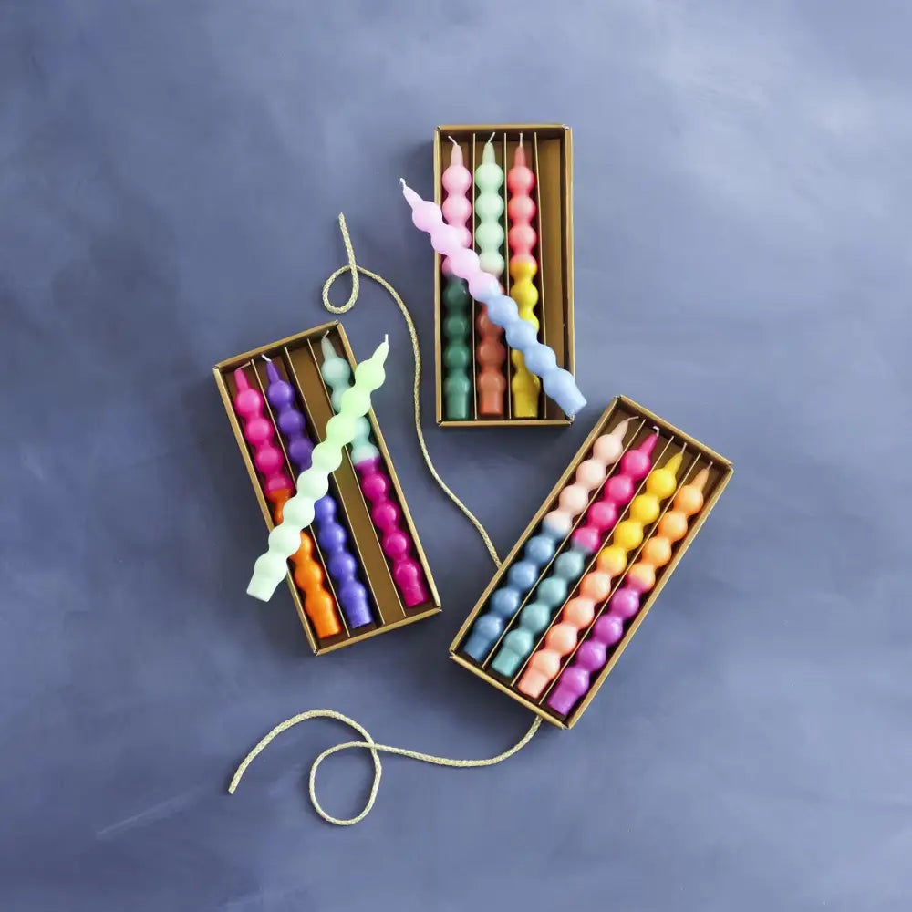 Three gold rectangular boxes containing colorful spiral candles, arranged on a textured slate blue background.