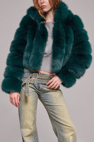 A woman wearing a cropped, voluminous emerald green faux fur jacket with horizontal banded panels, a wide lapel, and long banded sleeves, styled over a gray crop top and light-wash jeans.
