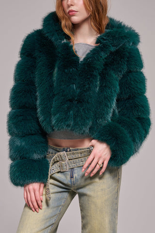A woman wearing a cropped, voluminous emerald green faux fur jacket with horizontal banded panels, a wide lapel, and long banded sleeves, styled over a gray crop top and light-wash jeans.