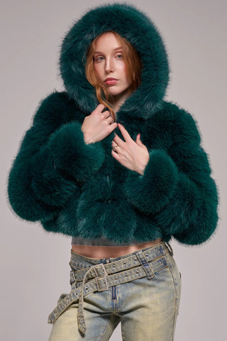 A woman wearing a cropped, voluminous emerald green faux fur jacket with horizontal banded panels, a wide lapel, and long banded sleeves, styled over a gray crop top and light-wash jeans.