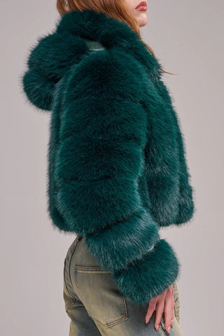 A woman wearing a cropped, voluminous emerald green faux fur jacket with horizontal banded panels, a wide lapel, and long banded sleeves, styled over a gray crop top and light-wash jeans.