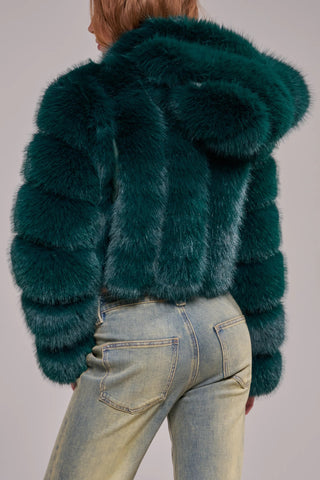 A woman wearing a cropped, voluminous emerald green faux fur jacket with horizontal banded panels, a wide lapel, and long banded sleeves, styled over a gray crop top and light-wash jeans.