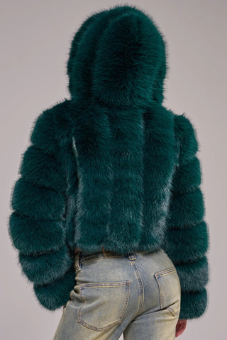 A woman wearing a cropped, voluminous emerald green faux fur jacket with horizontal banded panels, a wide lapel, and long banded sleeves, styled over a gray crop top and light-wash jeans.