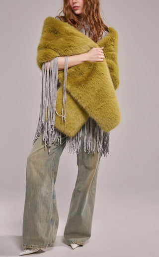 A model wearing an oversized plush faux fur wrap scarf in warm olive green with long gray yarn fringe trim, draped over both shoulders and paired with wide-leg jeans.