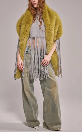 A model wearing an oversized plush faux fur wrap scarf in warm olive green with long gray yarn fringe trim, draped over both shoulders and paired with wide-leg jeans.