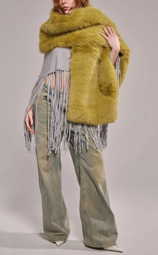 A model wearing an oversized plush faux fur wrap scarf in warm olive green with long gray yarn fringe trim, draped over both shoulders and paired with wide-leg jeans.