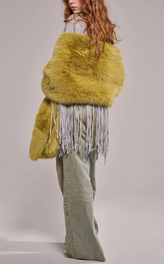 A model wearing an oversized plush faux fur wrap scarf in warm olive green with long gray yarn fringe trim, draped over both shoulders and paired with wide-leg jeans.