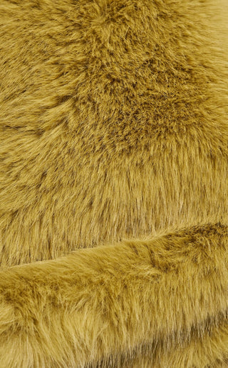A model wearing an oversized plush faux fur wrap scarf in warm olive green with long gray yarn fringe trim, draped over both shoulders and paired with wide-leg jeans.