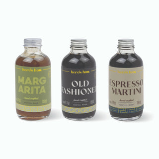 Three small glass cocktail mix bottles with silver caps on a white background: 'here's how' Marg Arita, Old Fashioned, and Espresso Martini.