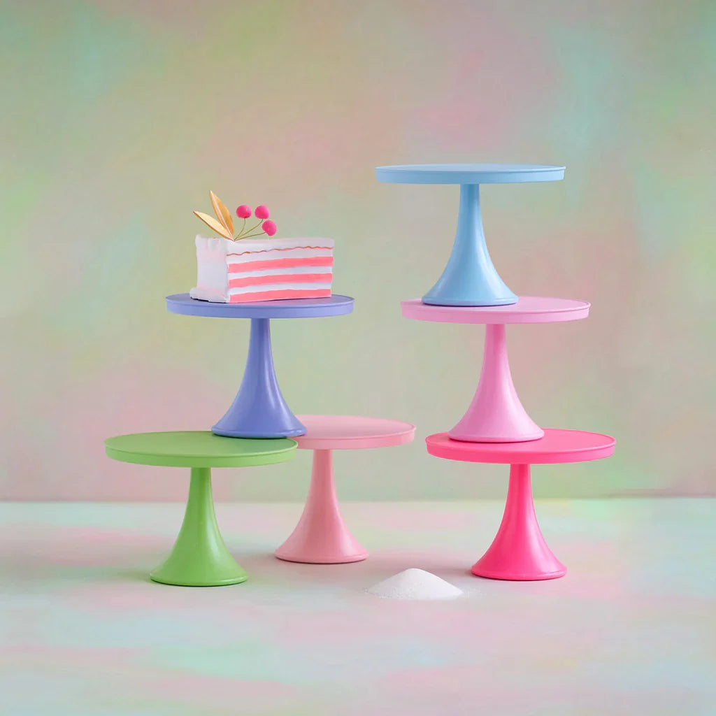 Four pastel-colored cake stands in pink, blue, purple, and green, arranged on a soft gradient background. One stand holds a small pink-and-white striped cake decoration.