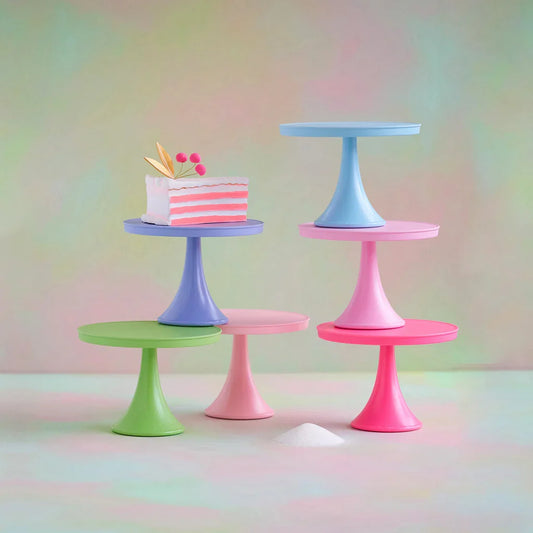 Four pastel-colored cake stands in pink, blue, purple, and green, arranged on a soft gradient background. One stand holds a small pink-and-white striped cake decoration.