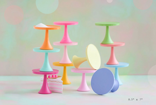 A set of pastel-colored miniature display stands and cake tiers, including pink, blue, green, orange, yellow, and purple pieces, arranged on a light surface against a soft pastel background with bokeh circles.