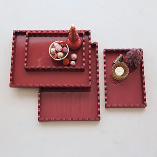 Three nested rectangular serving trays in a deep red color with a textured beaded border, styled with decorative objects on a light off-white background.
