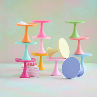 A set of pastel-colored miniature cake stands and display pedestals arranged on a soft pastel background. The set includes multiple stands in pink, blue, green, orange, yellow, and purple, with one standing next to a small pink-and-white striped cake decoration.
