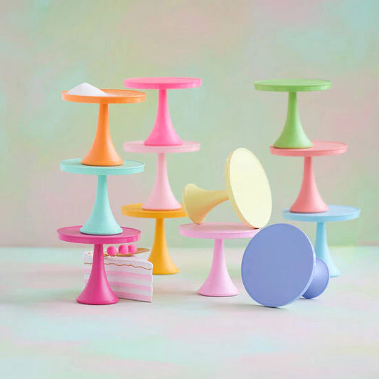 A set of pastel-colored miniature cake stands and display pedestals arranged on a soft pastel background. The set includes multiple stands in pink, blue, green, orange, yellow, and purple, with one standing next to a small pink-and-white striped cake decoration.