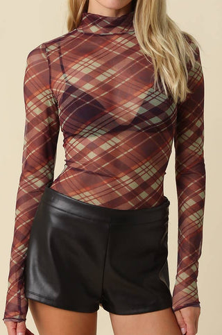 A woman modeling a long-sleeve mock neck top with a brown, red, and beige plaid pattern, paired with black faux leather shorts, against a solid beige background.