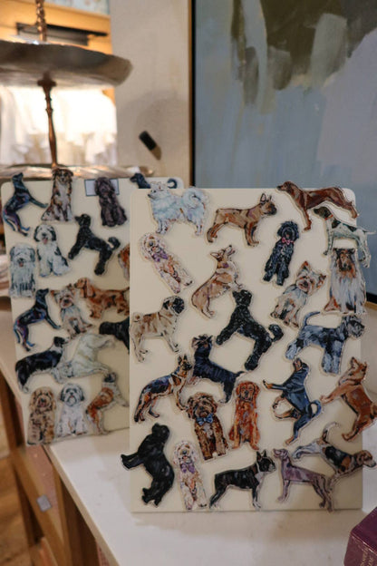 Two rectangular ceramic art pieces featuring hand-painted dog illustrations in various breeds and colors, displayed on a tabletop in a home or studio setting.