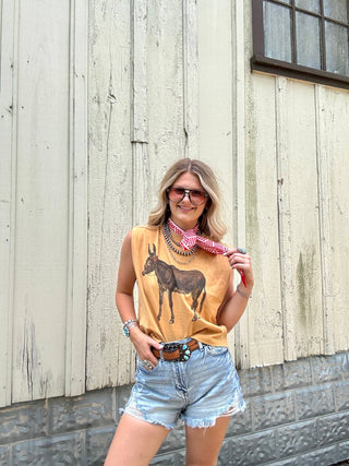 Woman wearing a mustard yellow tank top with a donkey graphic, denim shorts, and a red bandana, standing against a wooden wall.