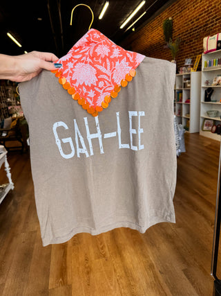 A person holds up a taupe-gray t-shirt with 'GAH-LEE' printed in white across the chest, paired with a coral-orange and white floral patterned bandana featuring orange scalloped edges.