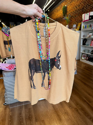 A sleeveless tan top held up by a hand in a boutique, featuring a graphic print of a black donkey and layered with colorful beaded necklaces.