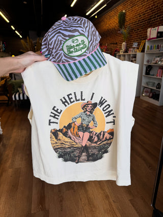 A sleeveless cream-colored graphic t-shirt held up by a hand inside a retail store, paired with a matching zebra-print and green striped baseball cap. The t-shirt features a vintage-style pin-up illustration of a woman in a desert landscape with the text 'THE HELL I WON'T'.