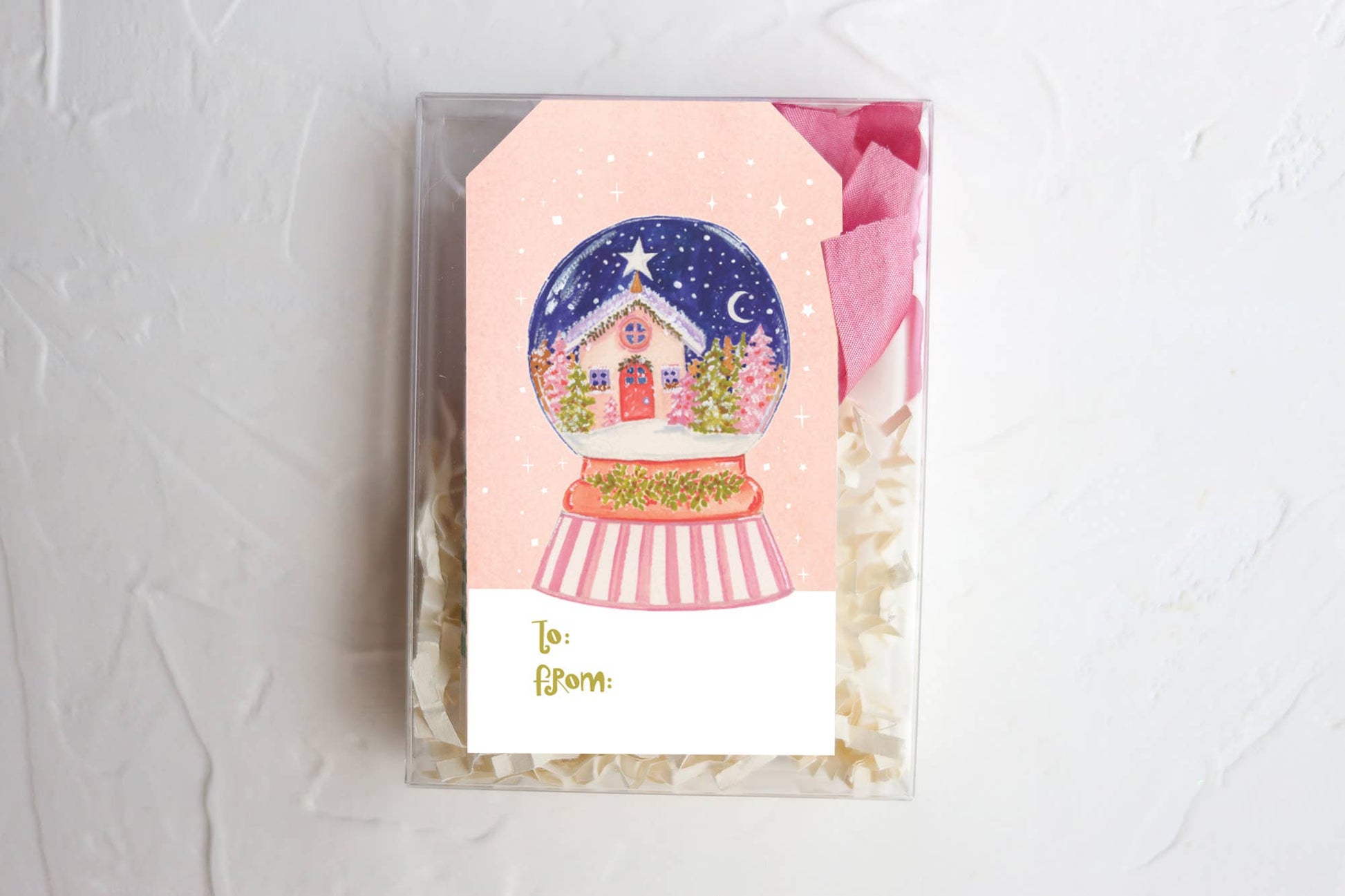 A pink gift tag with a snow globe design and a message 'To' on it, placed on a white surface, with a clear box packing visible behind it.